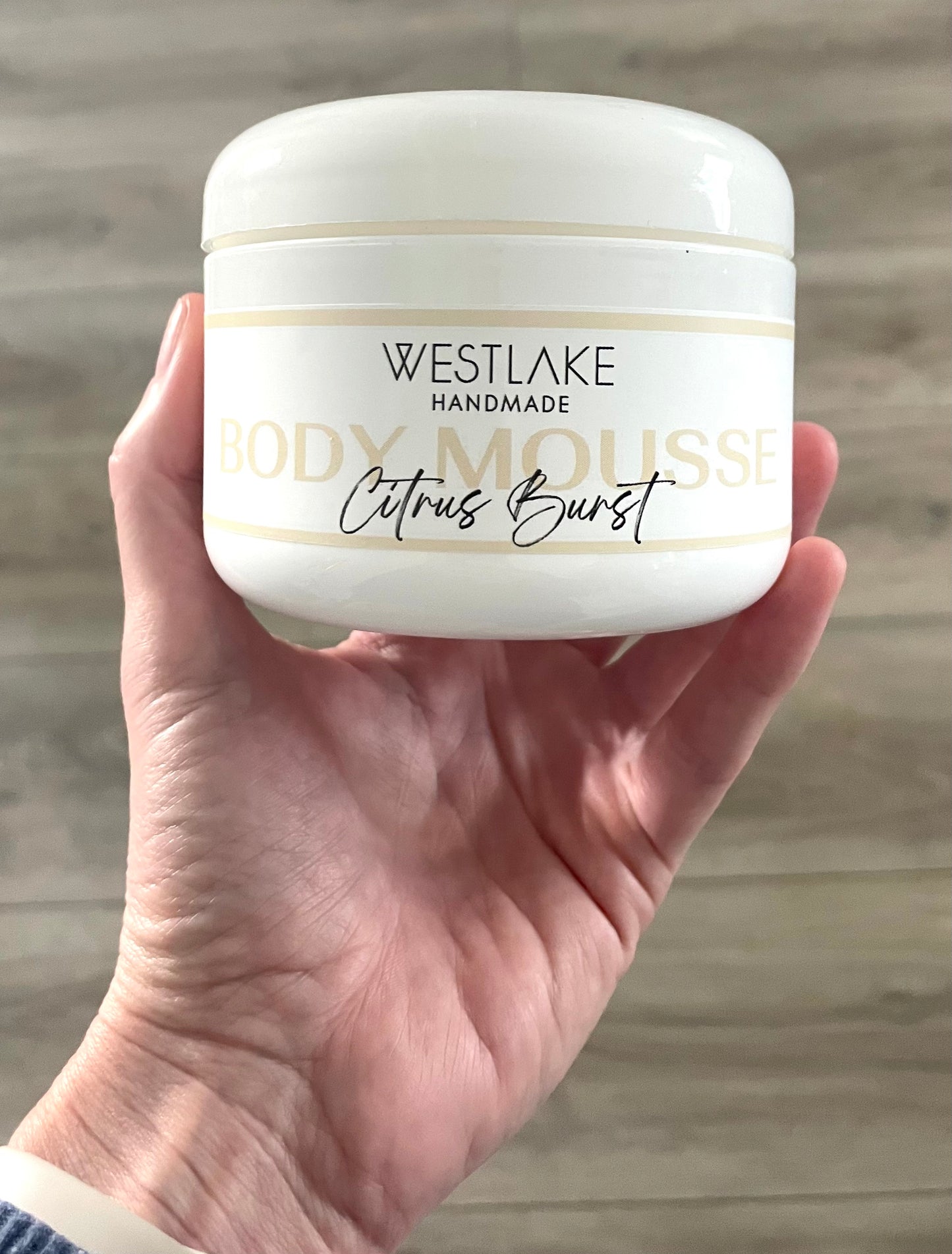 Hand holding a Westlake Handmade body mousse container with 'Citrus Burst' flavor on a wooden surface.