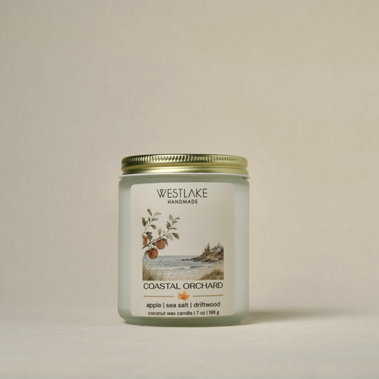 A Westlake Handmade coconut wax candle in a clear glass jar with a gold metal lid, featuring a white label with a coastal landscape illustration and the scent name 'Coastal Orchard'.