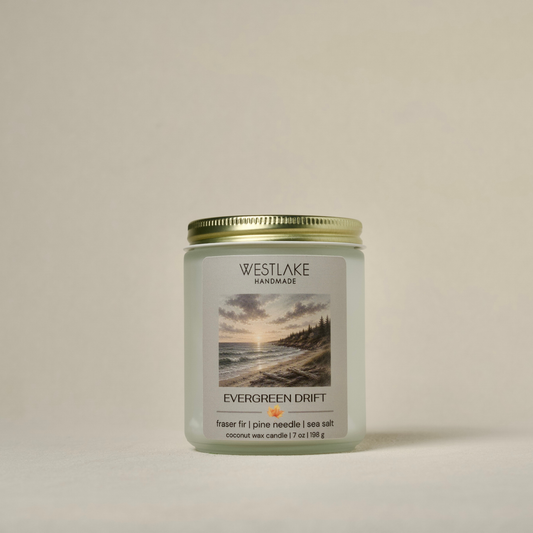 A Westlake Handmade coconut wax candle in a frosted glass jar with a gold metal lid. The front label features a scenic sunset beach illustration and the scent name 'Evergreen Drift'.