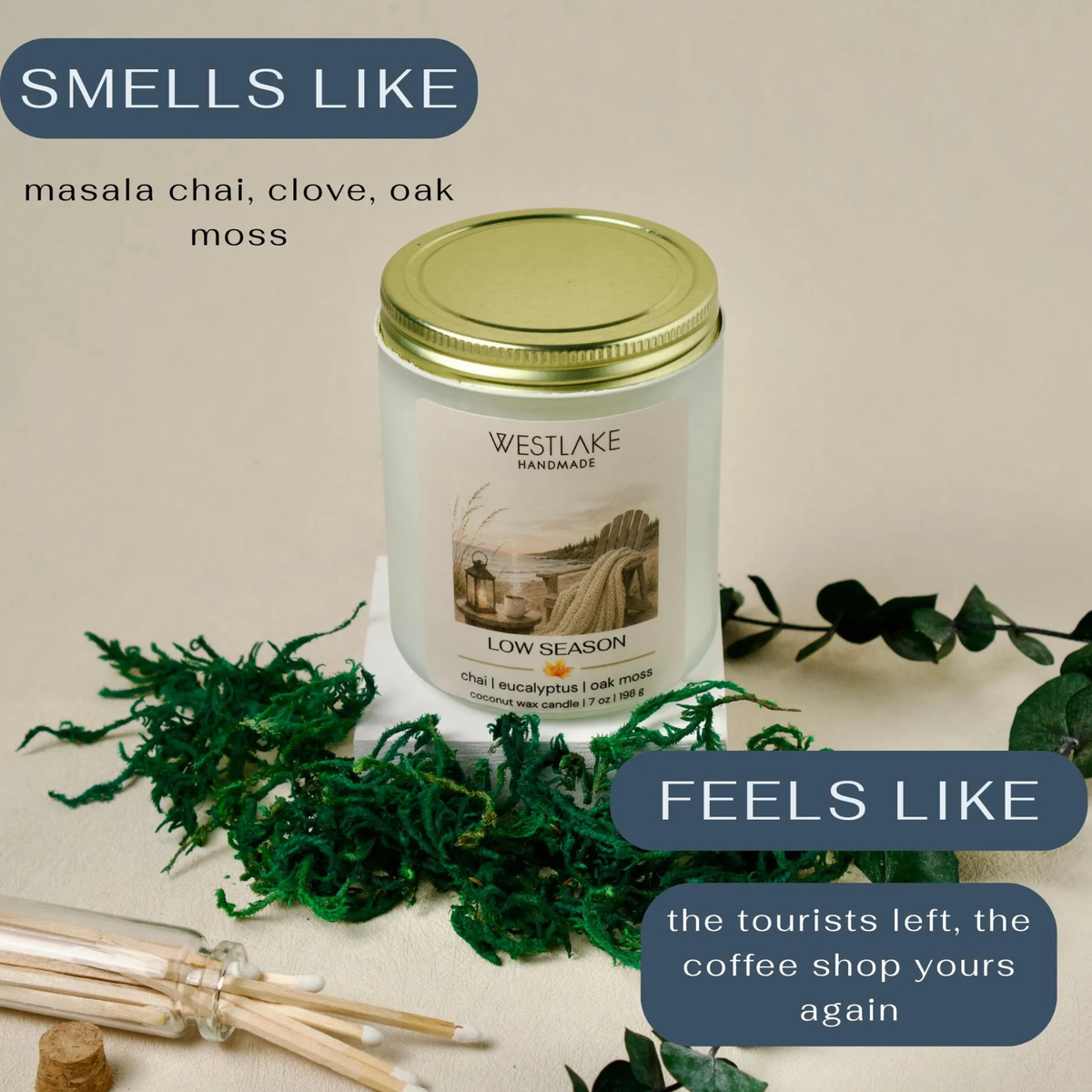 Westlake Handmade 'Low Season' soy candle in a frosted glass jar with a gold lid, styled with green moss, cotton wicks, and eucalyptus on a beige background with text overlays "smells ike" masala chai, clove, oak moss and "feels like" the tourists left, the coffee shop yours again.
