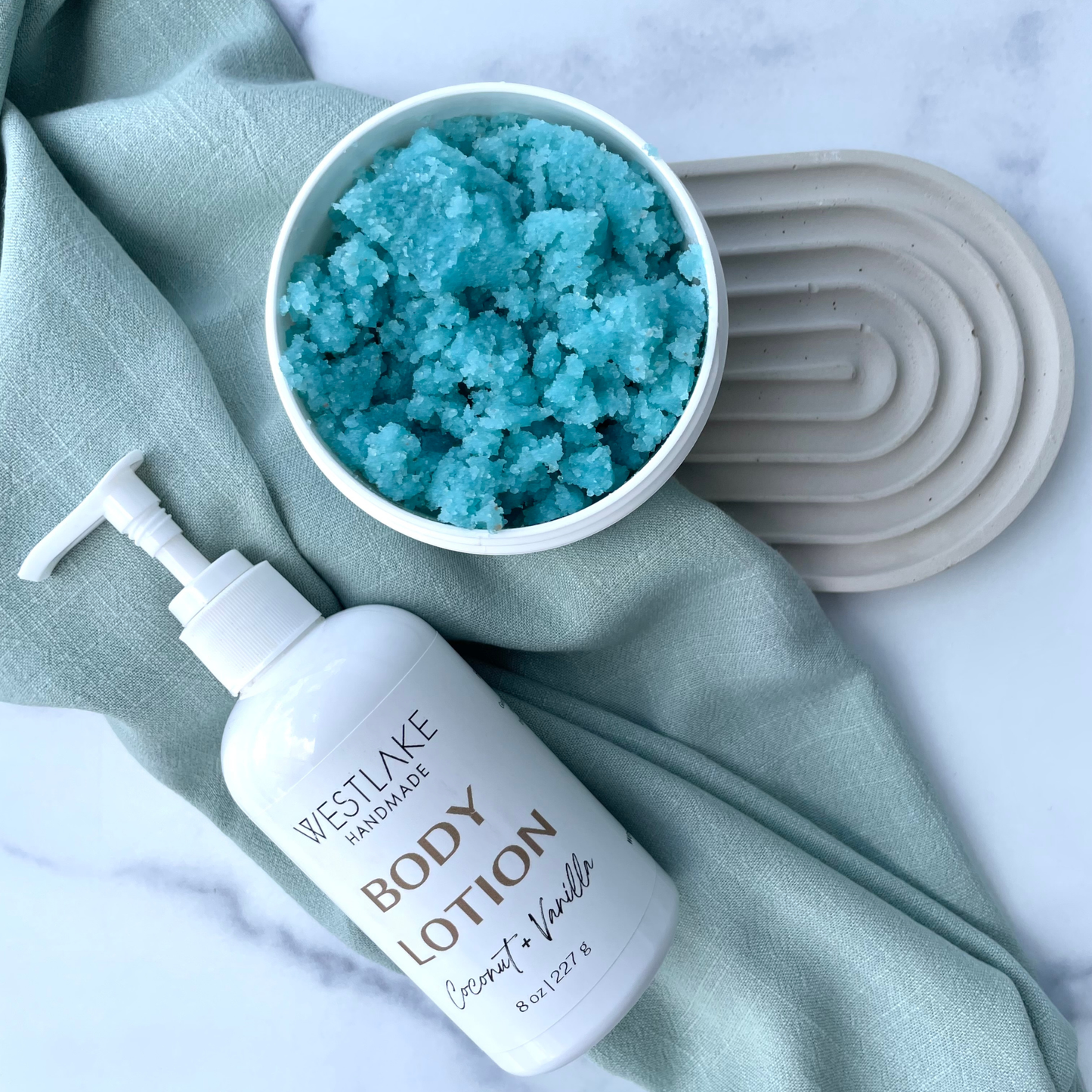 A bottle of Westlake Handmade Body Lotion in Coconut + Vanilla next to a white jar filled with bright blue salt scrub, styled on a sage green cloth over a white marble surface.