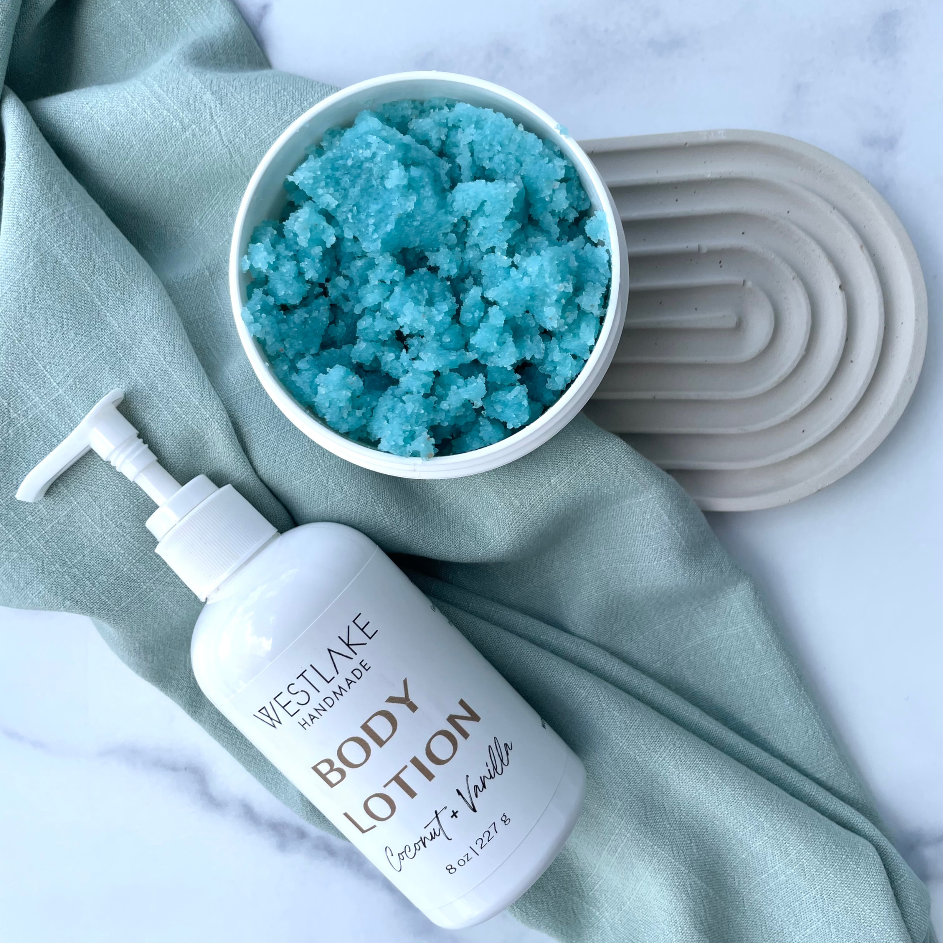 A bottle of Westlake Handmade Body Lotion in Coconut + Vanilla next to a white jar filled with bright blue salt scrub, styled on a sage green cloth over a white marble surface.