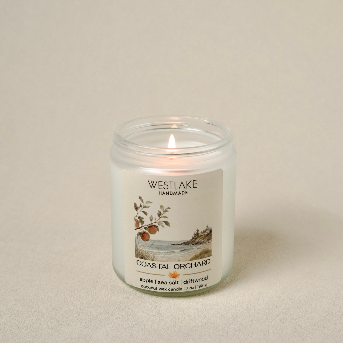 A lit Westlake Handmade Coastal Orchard scented candle in a clear glass jar, set against a neutral beige background.