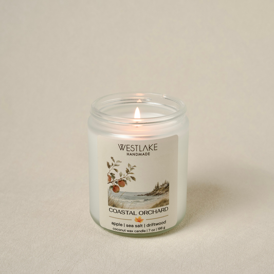 A lit Westlake Handmade Coastal Orchard scented candle in a clear glass jar, set against a neutral beige background.