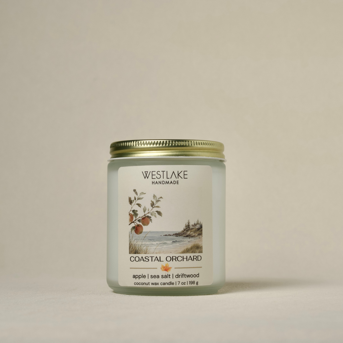 A Westlake Handmade coconut wax candle in a clear glass jar with a gold metal lid, featuring a white label with a coastal landscape illustration and the scent name 'Coastal Orchard'.