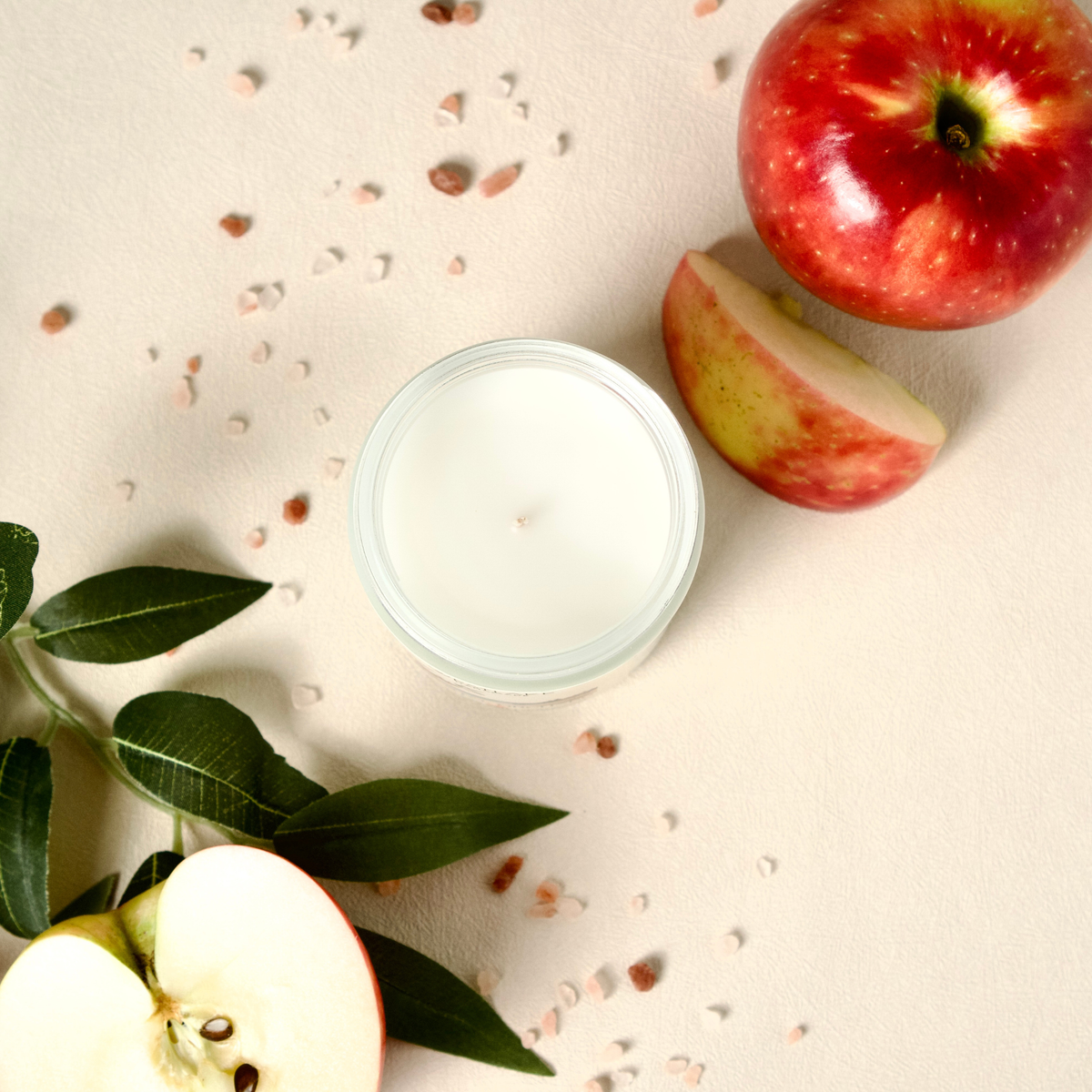 An open glass Westlake Handmade candle with white wax sits on a light beige surface, styled with red apples, apple slices, and green leaves with scattered decorative chips around it.