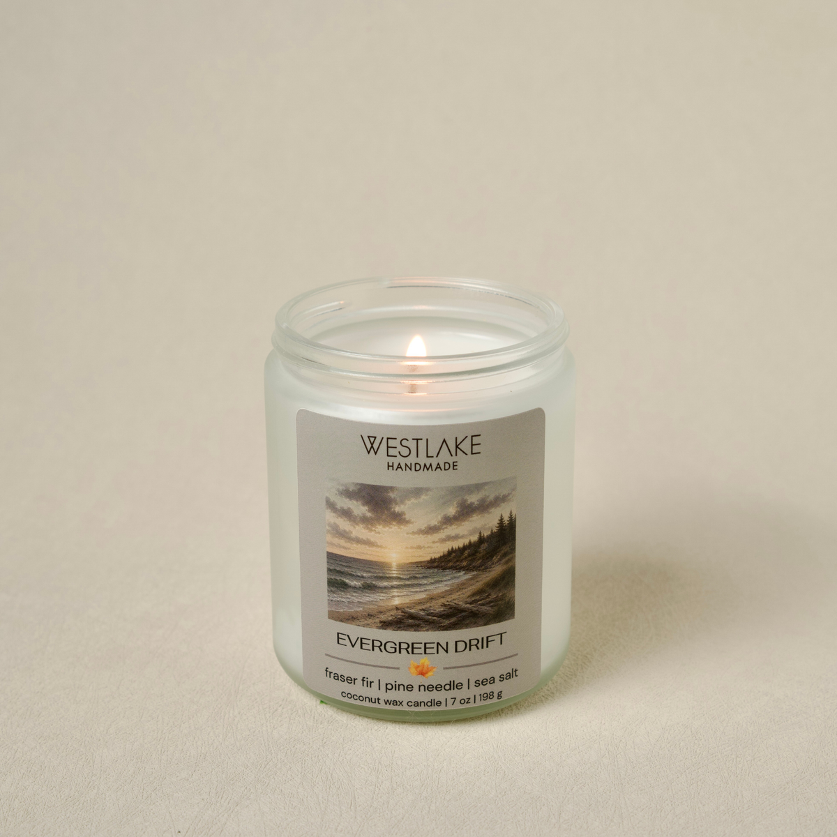 A lit Westlake Handmade wax candle in a clear glass jar with a white label reading 'Evergreen Drift' and a scenic landscape photo, set against a neutral beige background.