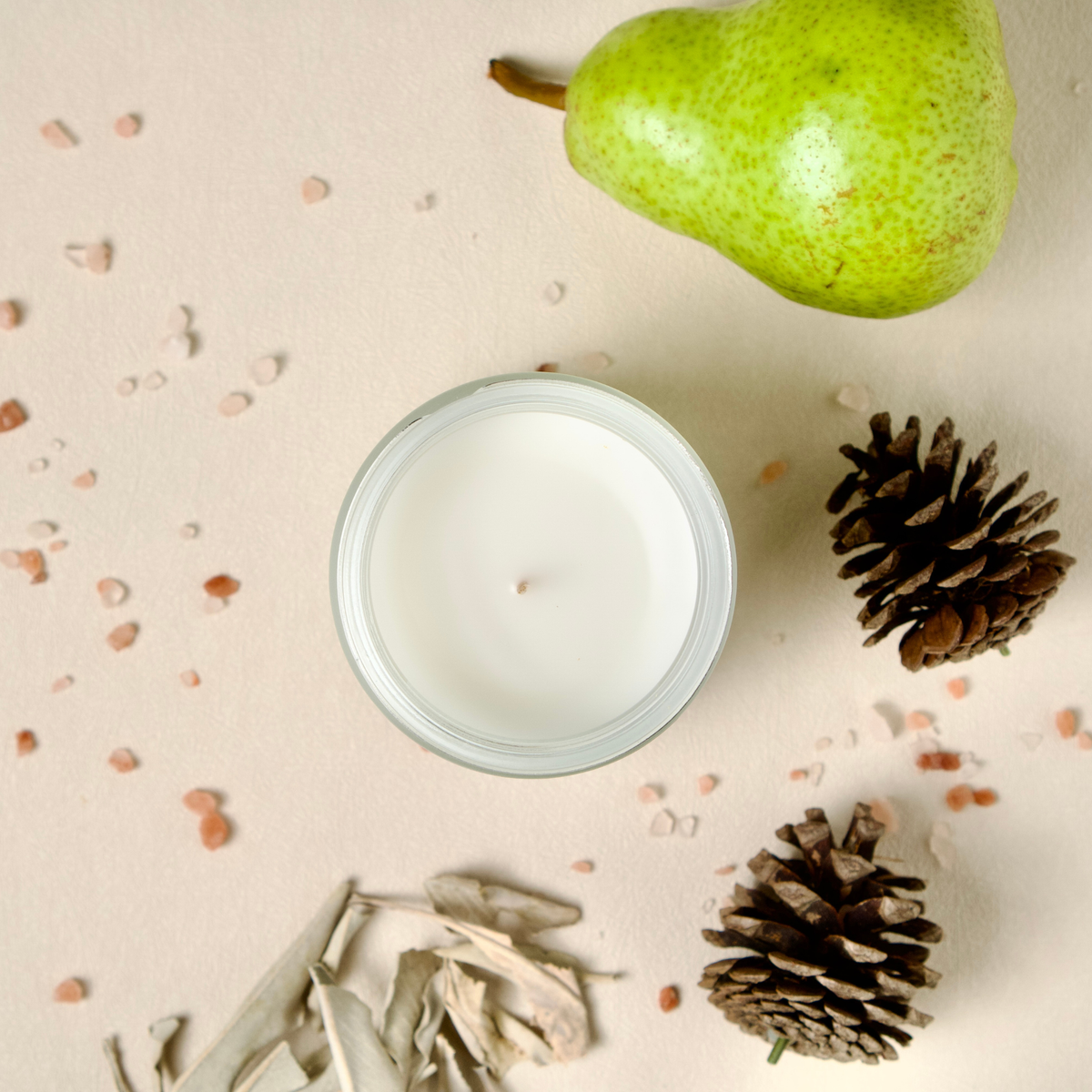 A Westlake Handmade candle in a clear glass jar is arranged on a light beige surface with a green pear, two pinecones, and dried leaves around it.