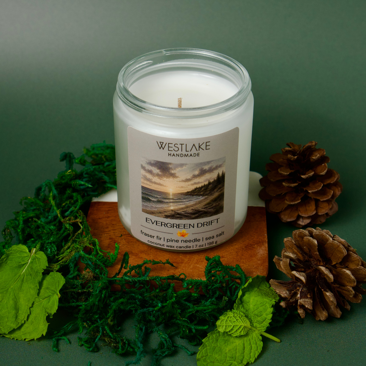 A Westlake Handmade candle in a clear glass jar with a printed label reading Westlake Handmade Evergreen Drift, styled on a wooden block with green moss, pinecones, and leaves against a dark green background.