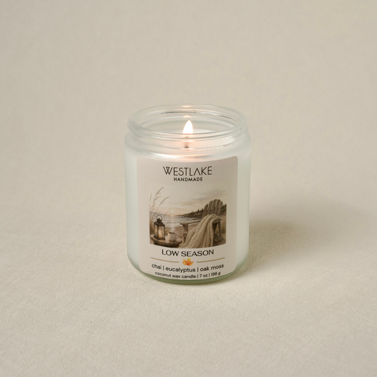 A lit coconut wax candle in a clear glass jar with a white label reading 'WESTLAKE HANDMADE' and 'LOW SEASON', set against a neutral beige background.