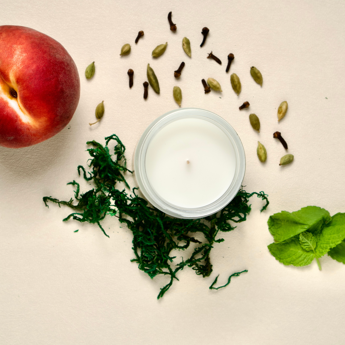 A Westlake Handmade candle in a clear glass jar, styled in a flat lay with a peach, green moss, cardamom seeds, and mint leaves on a light beige background.