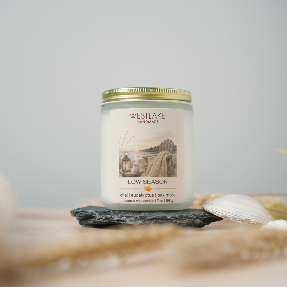 A Westlake Handmade coconut wax candle in a clear glass jar with a gold metal lid, labeled 'Low Season'. The candle is styled on a dark slate stone with a white seashell and dried grass against a soft gray background.