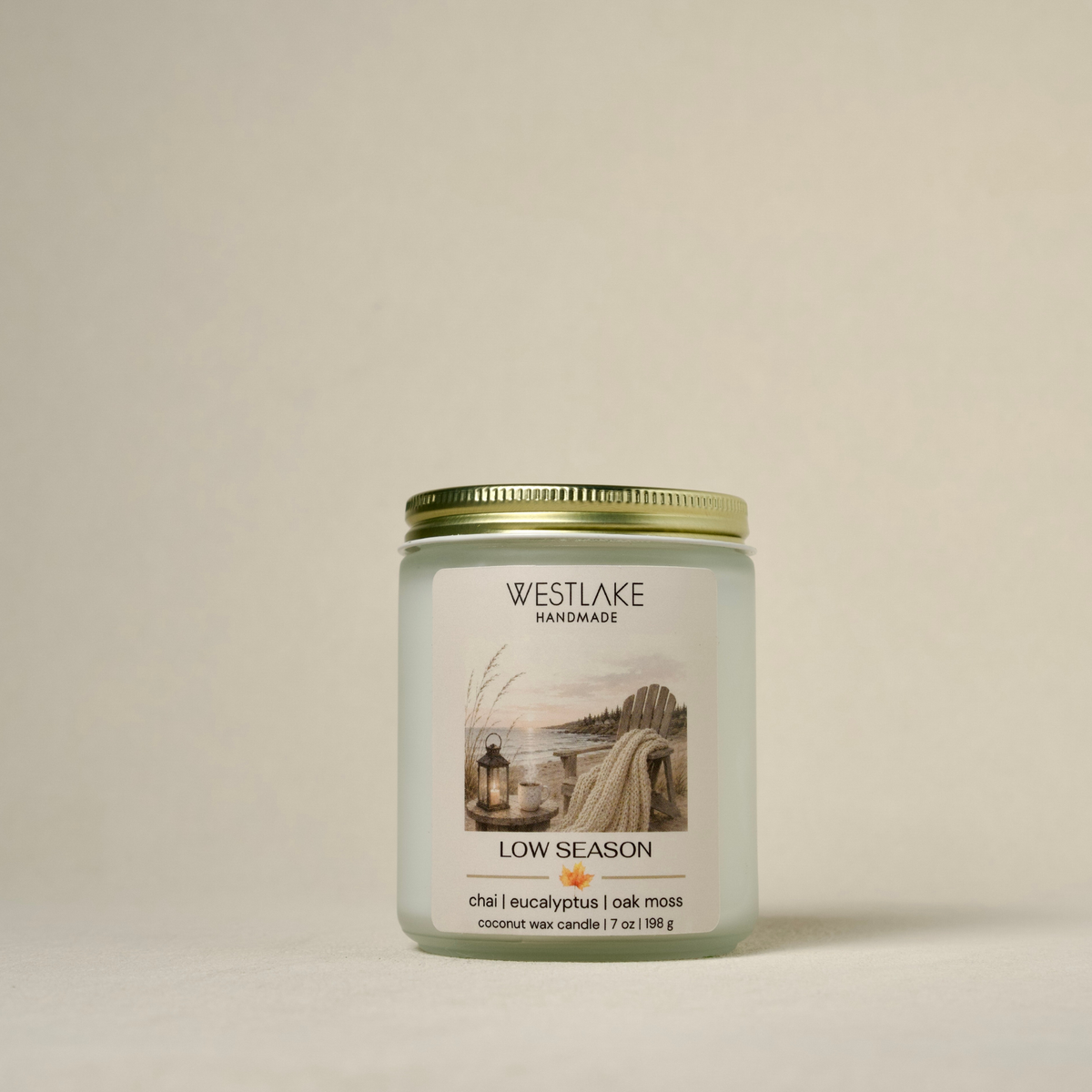 A Westlake Handmade 'Low Season' coconut wax candle in a frosted glass jar with a gold metal lid, set against a solid, warm off-white background.