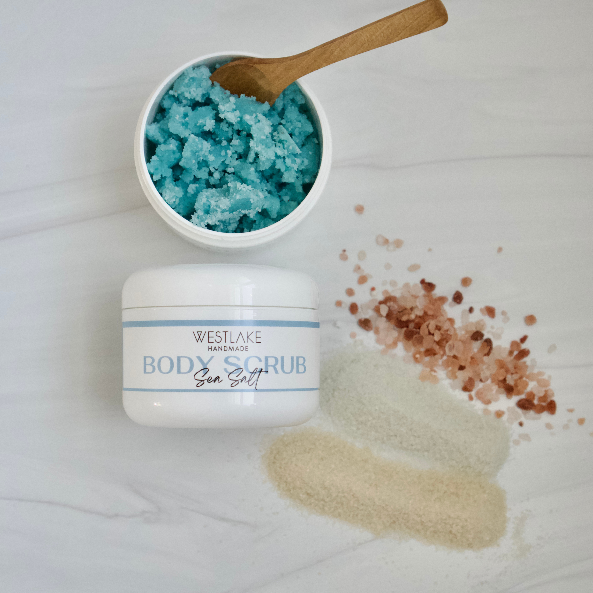 Westlake Handmade Sea Salt body scrub jar with a white lid, shown next to an open jar filled with bright blue scrub and a wooden spoon, on a light marble surface with scattered pink and beige salt crystals.