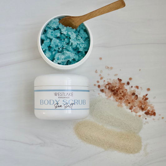 Westlake Handmade Sea Salt body scrub jar with a white lid, shown next to an open jar filled with bright blue scrub and a wooden spoon, on a light marble surface with scattered pink and beige salt crystals.