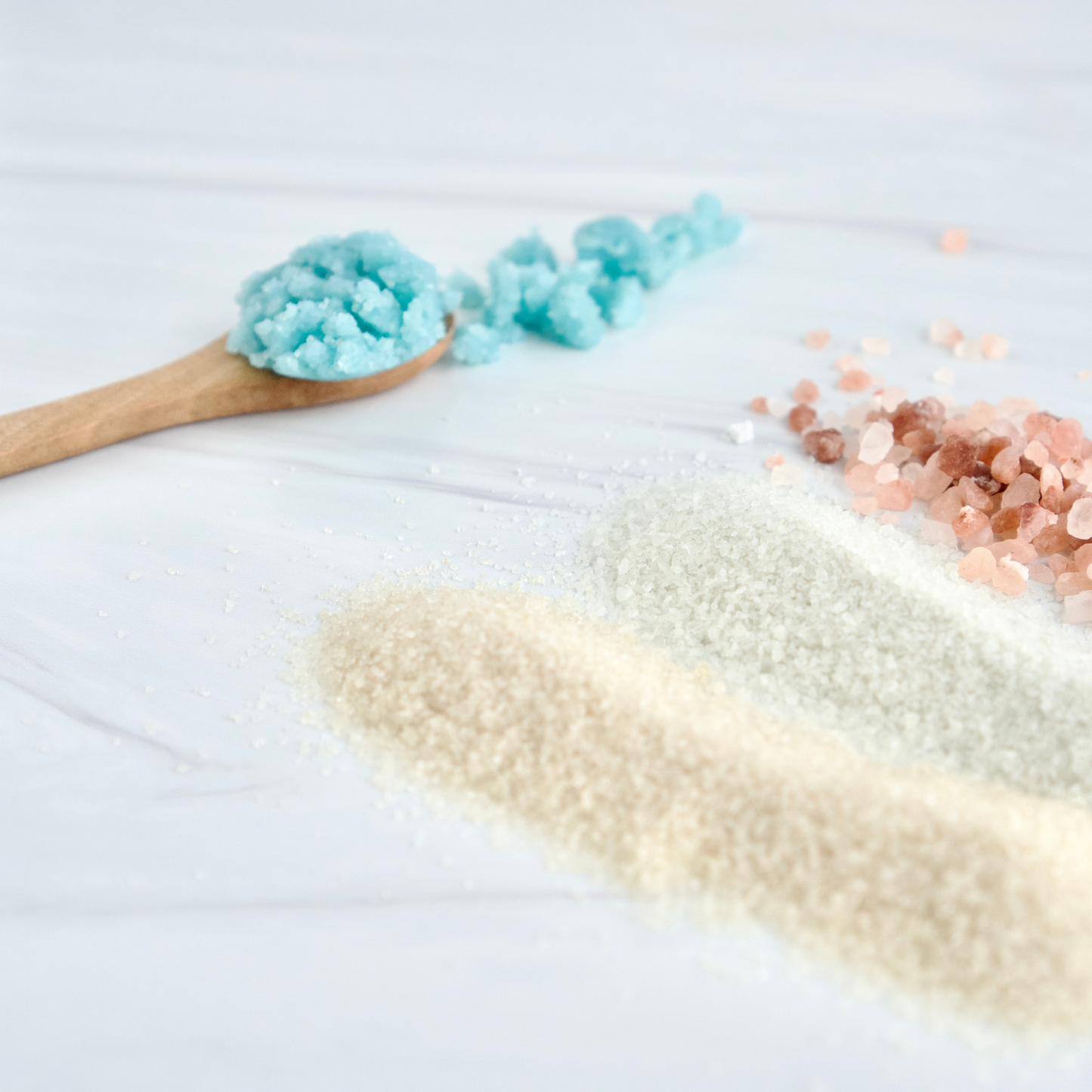 Three types of exfoliants arranged on a white surface: a wooden spoon holding bright blue salt scrub. A pile of pale brown sugar, a pile of pale green salts, and a small pile of pink rose-gold salts.
