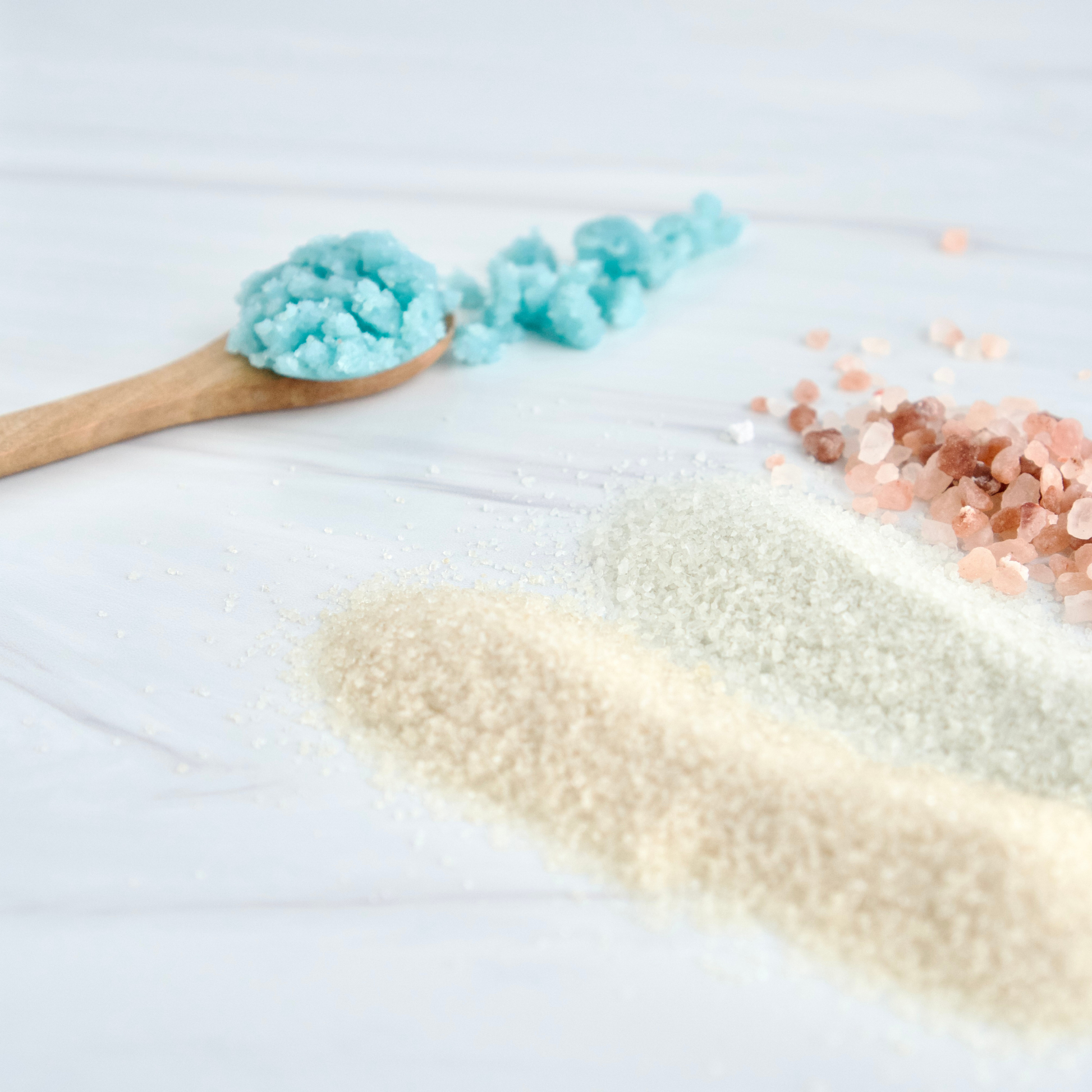 Three types of exfoliants arranged on a white surface: a wooden spoon holding bright blue salt scrub. A pile of pale brown sugar, a pile of pale green salts, and a small pile of pink rose-gold salts.