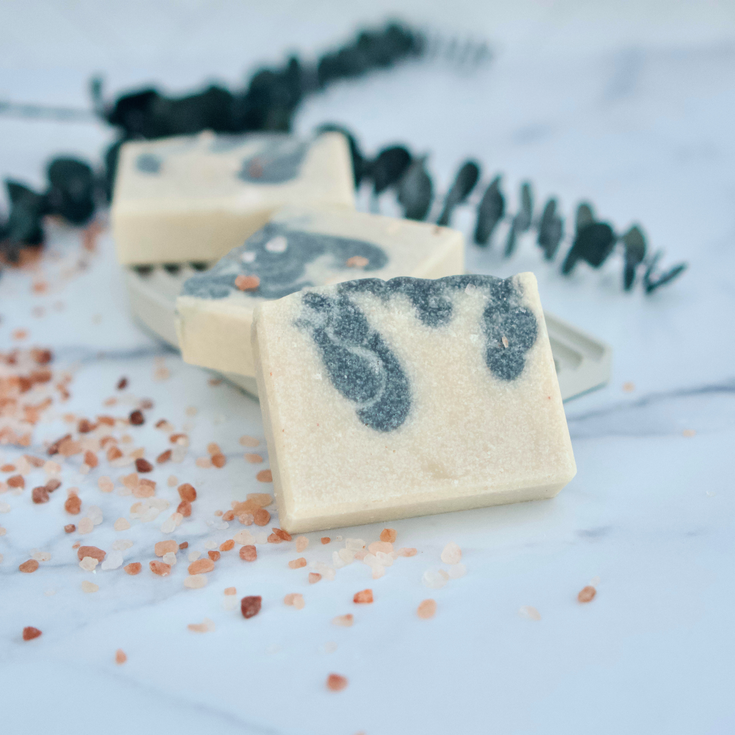 Three bars of soap with gray exfoliating scrub on a light surface with pink Himalayan salt.