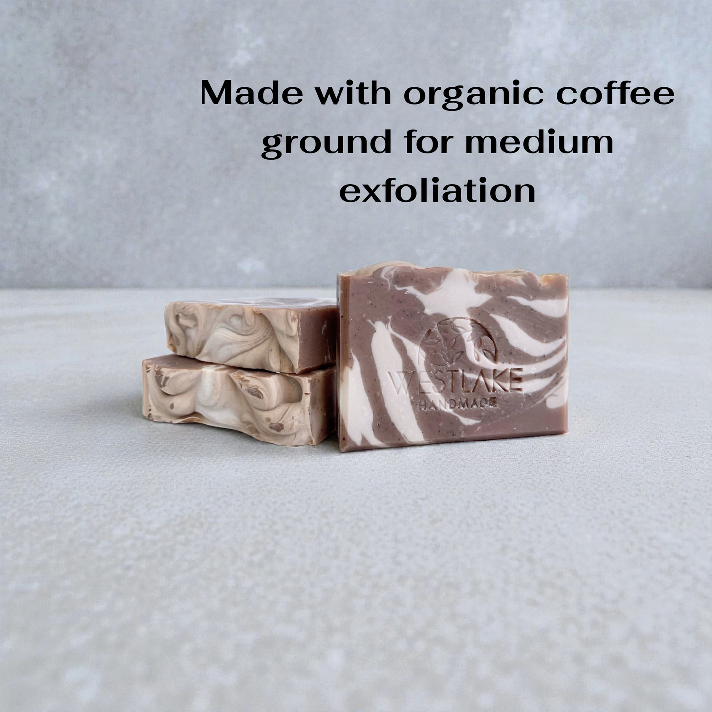 Three marbled coffee-scented soap bars on a textured surface with text about organic coffee ground exfoliation.